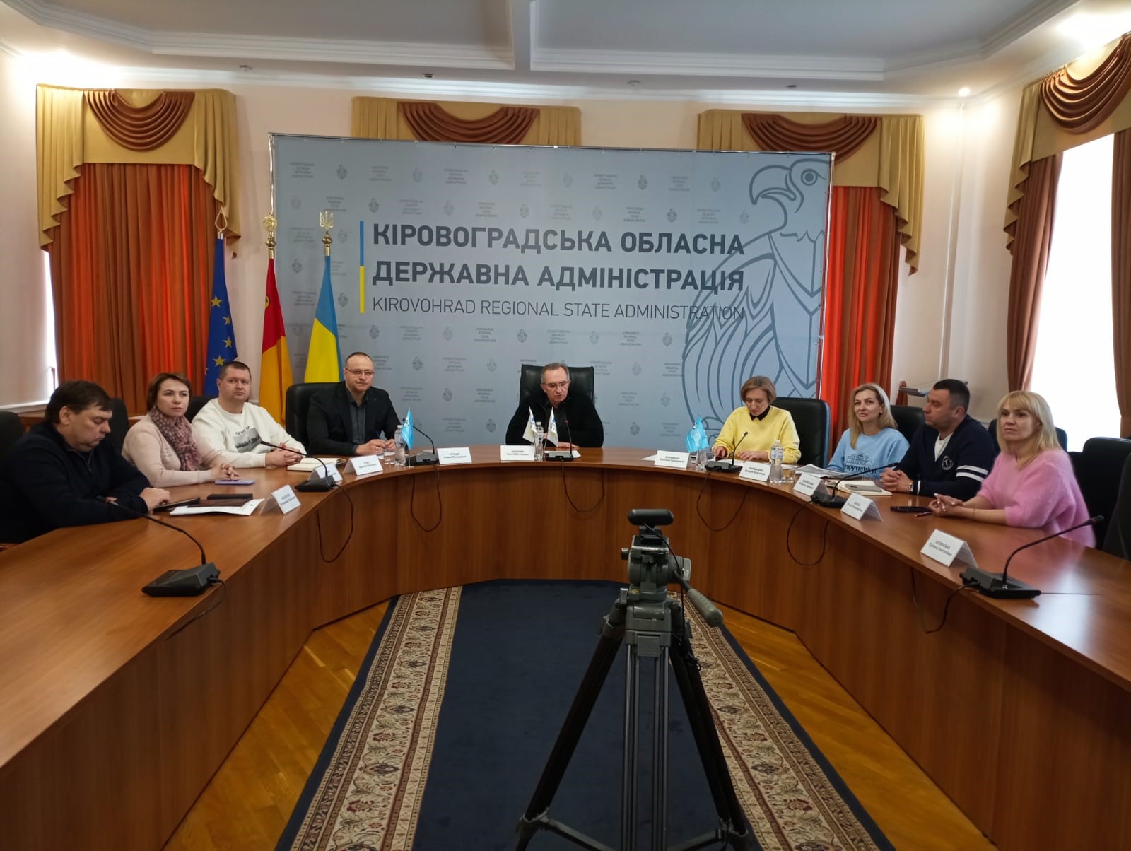 Офіційна зустріч керівництва Кіровоградської обласної державної адміністрації за круглим столом у конференц-залі з прапорами та відеокамерою на передньому плані.
